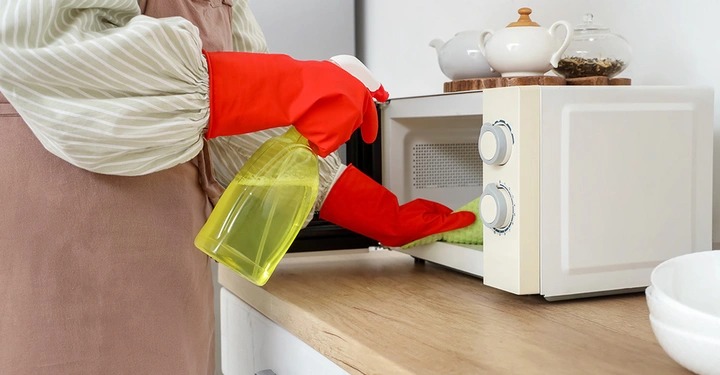 Eine Person trägt knallrote Putzhandschuhe und putzt ihre Mikrowelle mit einem Mikrofasertuch un speziellem Putzmittel.