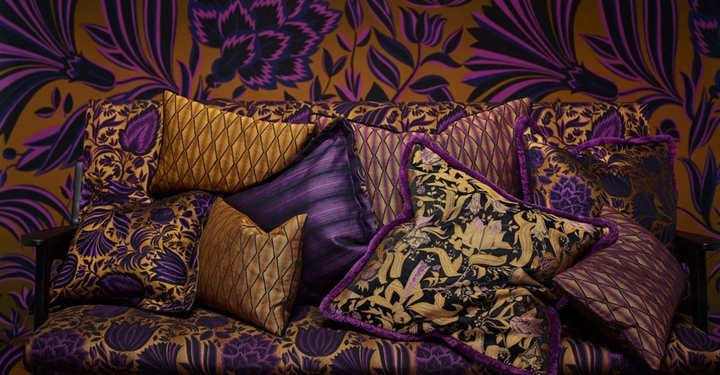 Mehrere dekorative Kissen mit goldenen und violetten Mustern liegen auf einem passenden stoffbezogenen Sofa in einem Raum mit gemusterten Tapeten in denselben Farben.