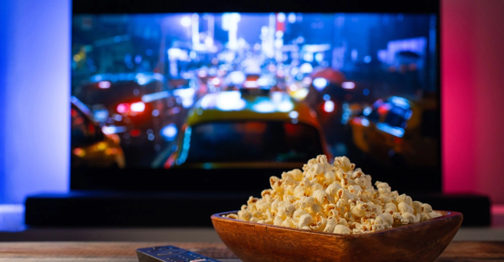 Popcorn in schöner Holzschale steht auf Fernsehtischchen. Im Hintergrund ein großer Bildschirm, auf dem ein Film läuft. 