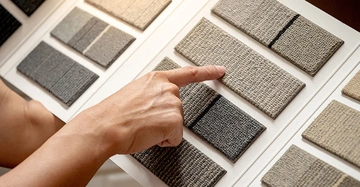 Person deutet mit ausgestreckter Hand auf ein Brett mit verschiedenen Mustern von Teppichböden.