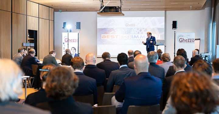 Galerie-Bild 3: Ein Mann hält eine Präsentation vor einem sitzenden Publikum in einem Konferenzraum. Auf dem Bildschirm hinter ihm steht Ghezzo BESTandIMMO17. Das Publikum hört aufmerksam zu und der Raum hat Holzverkleidungen.
