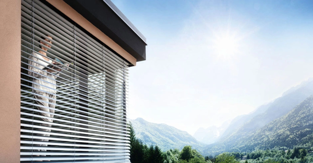 Frau steht vor halbgeöffnetem Raffstore und sieht entspannt auf die idyllische Berglandschaft vor ihrem Haus.