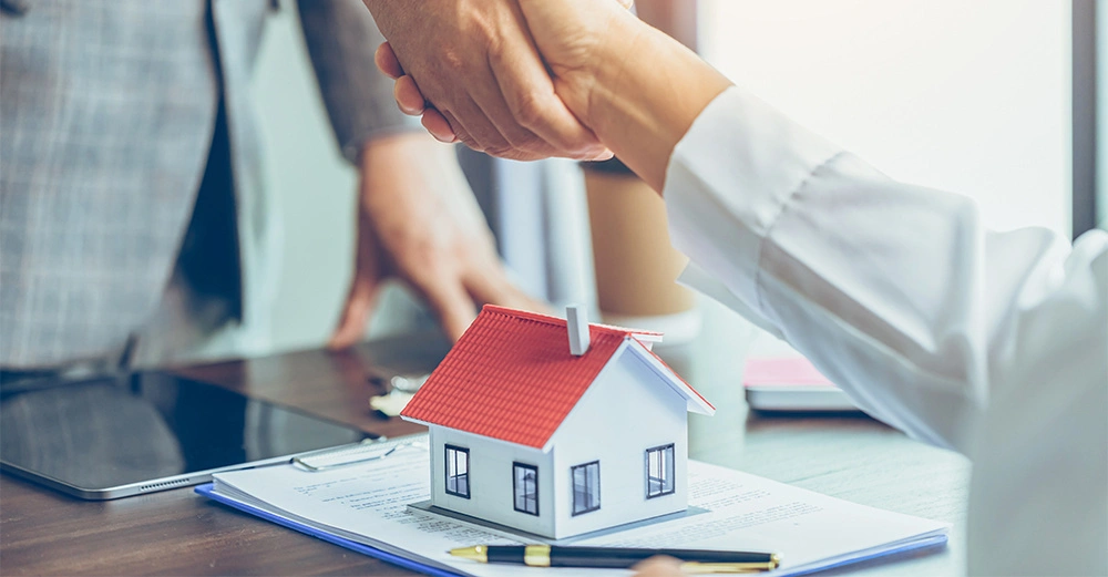 Miniaturhaus steht auf Dokumenten zwei Menschen schütteln Hände Laptop Stift im Bürohintergrund