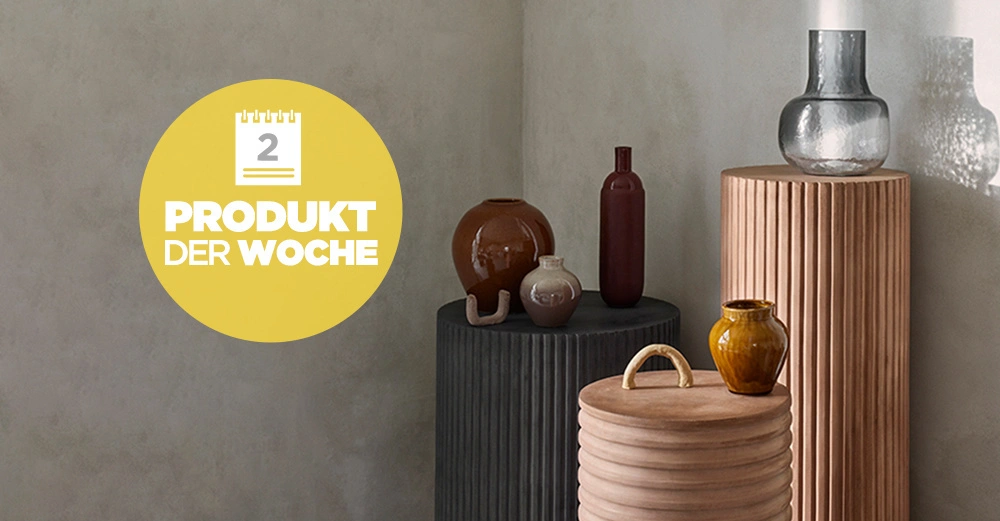 Vasen stehen auf wellenförmigen Podesten in einem minimalistischen Raum. Gelbes Plakat mit Kalender und Text PRODUKT DER WOCHE 2 links in der Bildmitte.