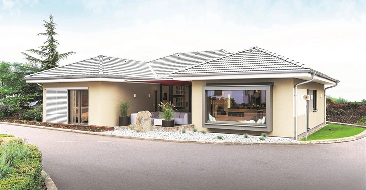 Ein modernes, einstöckiges Haus mit hellem Putz und weißen Dachziegeln steht auf einem gepflasterten Weg; drumherum gepflegte Gärten mit Sträuchern und Rasen.