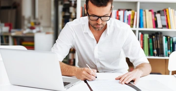 Ein Mann sitzt an einem Schreibtisch und schreibt auf ein Dokument; neben ihm steht ein offener Laptop; im Hintergrund befinden sich Bücherregale in einem Bürozimmer.