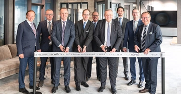 Gruppenfoto: Neun Männer im Anzug von Alukönigstahl und Schüco hinter dem Eröffnungsband. Drei davon mit Schere, um das Band durchzuschneiden.