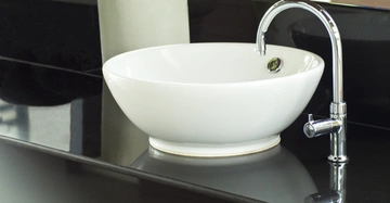 Ein weißes Waschbecken ist auf einer glänzenden schwarzen Arbeitsfläche installiert mit einem silbernen Wasserhahn der darüber an einer Wand montiert ist.