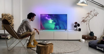 Ein Mann sitzt auf einem Stuhl und schaut auf einen eingeschalteten Fernseher in einem modern eingerichteten Wohnzimmer mit Holzscheiten, einem Teppich und einer Stehlampe.