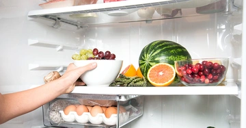 Eine Hand platziert eine Schüssel mit grünen und roten Trauben in einem Kühlschrank der mit verschiedenen Früchten wie Wassermelone, Orange, Preiselbeeren und Eiern gefüllt ist.