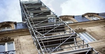 Gerüst ragt senkrecht in die Höhe ein beigefarbenes historisches Gebäude mit großen Fenstern und schmiedeeisernen Balkonen ist im Hintergrund zu sehen der Himmel ist bewölkt