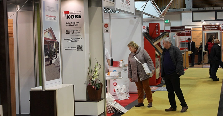 Galerie-Bild 16: Zwei Personen gehen durch eine Messehalle mit verschiedenen Ständen. Ein Stand zeigt die Firmeninformationen: Markisen-Station KOBE Gallenberg 17b 4950 Altheim Oberlangstraßl 3 4921 Hohenzell An der Fernstraße 2 4770 Andorf Wohnnet Messe Halle 6 130.