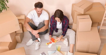 Ein Mann und eine Frau studieren Farbmuster während sie auf dem Boden zwischen vielen Umzugskartons sitzen in einem Raum mit Holzboden und einem Pflanzenabschnitt links.