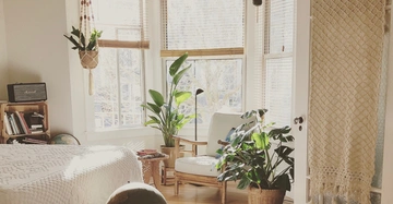 Ein gemütlicher Raum mit einem weißen Bett eine Holzkommode mit Büchern und einem Radio und mehreren Pflanzen auf dem Boden und an den Fenstern daneben ein bequemer Sessel im Sonnenlicht.