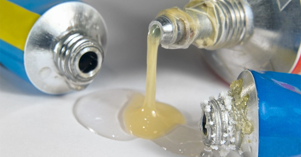 Drei geöffnete Farbtuben liegen auf einer Oberfläche während gelbliche und klare Flüssigkeit ausläuft umgeben von einer hellen Umgebung.