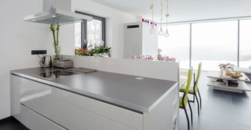 Moderne Küche mit grauer Arbeitsplatte und Induktionsherd. Blumenvasen und Toaster auf der Arbeitsplatte. Essbereich mit grünen Stühlen und großem Fenster im Hintergrund, daneben ein kleiner Tisch.