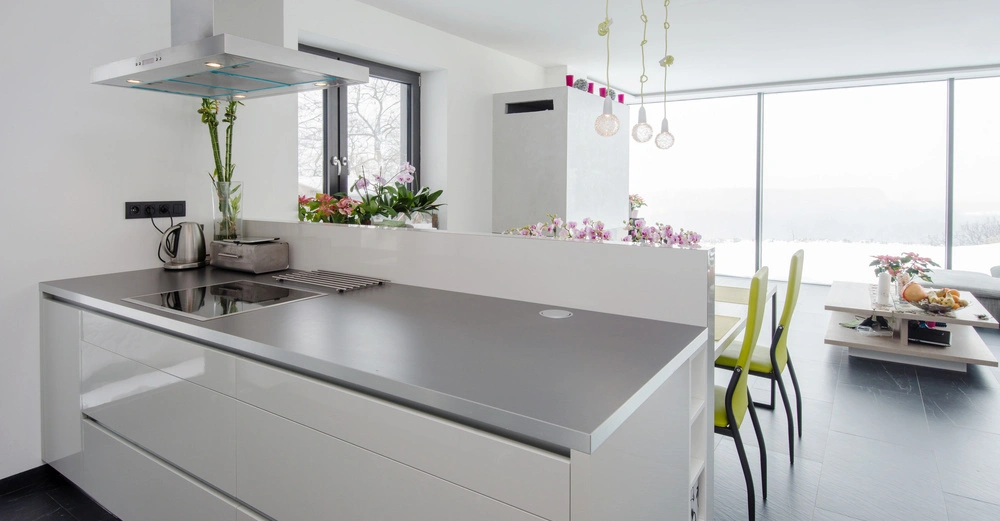 Moderne Küche mit grauer Arbeitsplatte und Induktionsherd. Blumenvasen und Toaster auf der Arbeitsplatte. Essbereich mit grünen Stühlen und großem Fenster im Hintergrund, daneben ein kleiner Tisch.