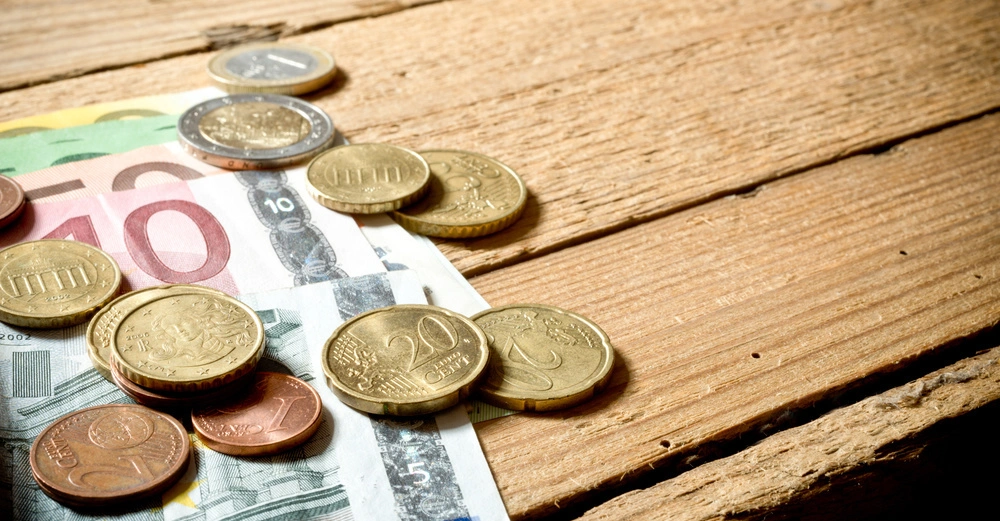 Münzen und Euro-Banknoten liegen verstreut auf einem rustikalen Holztisch. Mehrere Gold- und Kupfermünzen verschiedener Wertigkeiten sowie Banknoten in 10- und 100-Euro-Scheinen.