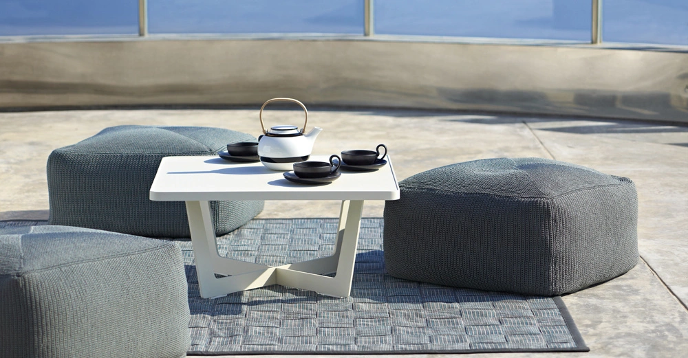 Tisch mit Teeset darauf steht auf einem Teppich und zwischen drei gepolsterten Hockern auf einer großen Terrasse im Freien.