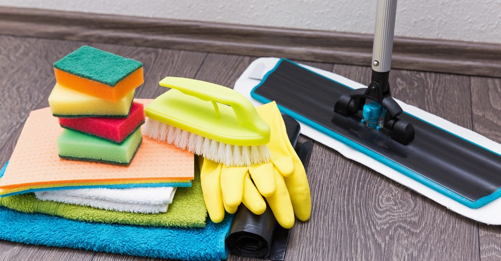 Dunkler Laminatboden mit einem Wischmopp neben einem bunten Haufen aus Wischtüchern und Putzschwämmen.