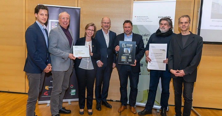 Sieben Personen stehen in einer Reihe und halten Zertifikate und ein Poster in den Händen in einem Konferenzraum. Text: "klimaaktiv" "klimaaktiv Bauen & Sanieren" "ÖGNB" "klimaaktiv Bronze"