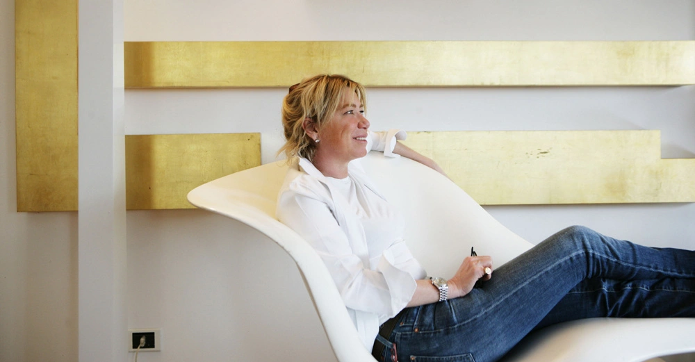 Frau sitzt entspannt auf einem modernen weißen Stuhl hält einen Stift lächelt und blickt zur Seite im Hintergrund weiße Wand mit horizontalen goldenen Streifen.