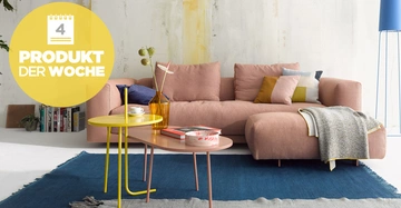 Rosa Sofa steht auf blauem Teppich mit bunten Kissen und Decke darin. Daneben zwei Tische mit Vase Buch und Tasse. Links gelbes rundes Label mit Text 4 PRODUKT DER WOCHE.