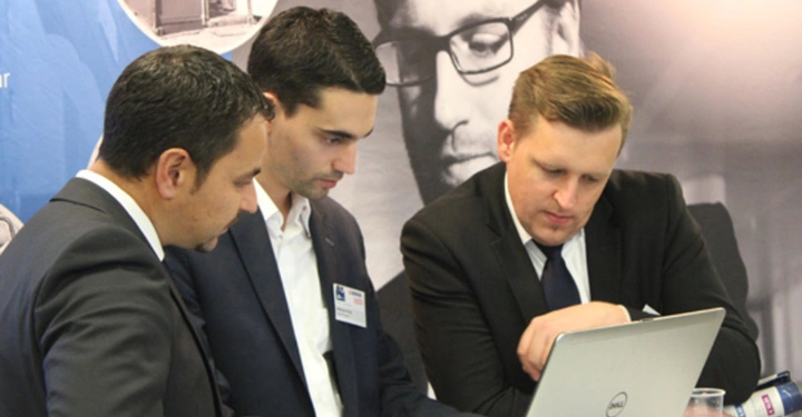 Galerie-Bild 10: Drei Männer in Anzügen betrachten gemeinsam einen Laptop in einem Geschäftsumfeld mit Postern an der Wand im Hintergrund. Einer zeigt auf den Bildschirm.