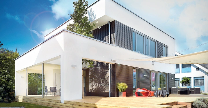 Perspektivische Aufnahme von weißem, zweistöckigem Fertighaus mit Flachdach und Dachterrasse sowie großer Fenstergfront. Blauer Himmel und Sonnenschein. 