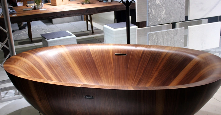 Galerie-Bild 15: Holzbadewanne steht auf Fliesenboden umgeben von modernen Möbeln und Dekorationen in einem Ausstellungsraum. Im Hintergrund stehen Tische mit Blumenvasen und Steinproben an der Wand. Text: Legno.