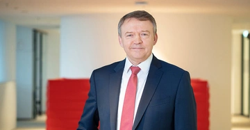 Mann im Anzug mit roter Krawatte steht lächelnd in einem modernen, hellen Büro mit roten Möbeln im Hintergrund.