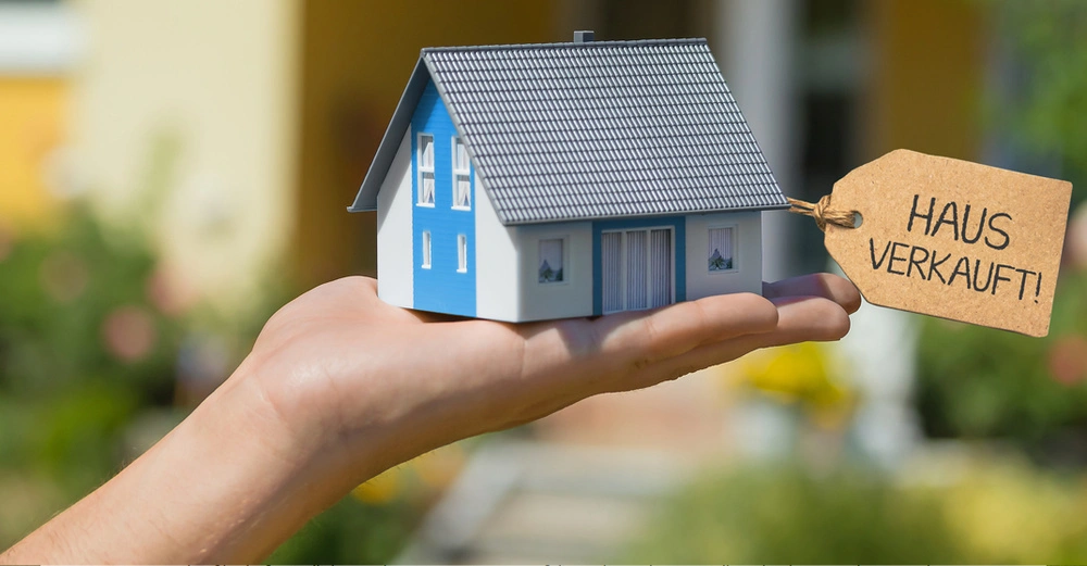 Person hält ein kleines Modellhaus mit einem Etikett in der Hand Inschrift auf dem Etikett HAUS VERKAUFT im Hintergrund ein unscharfer, grüner Garten.