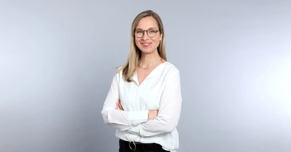 Eine Frau trägt eine Brille und lächelt mit verschränkten Armen vor einem hellgrauen Hintergrund. Sie trägt eine weiße Bluse und schwarze Hose.