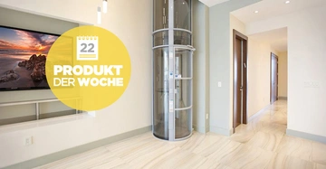 Wohnzimmer mit transparentem, runden Aufzug und großem an der Wand montierten Fernseher links. Gelber Kreis zeigt Text mit Kalender: 22 PRODUKT DER WOCHE. Modern eingerichtetes Haus mit Marmorfußboden.