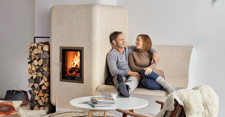 Galerie-Bild 5: Ein Paar sitzt lachend auf einer gefliesten Bank neben einem brennenden Kamin in einem gemütlichen Wohnzimmer, mit einem Holzstapel und einem Tisch mit Zeitschriften und einer Tasse im Vordergrund.