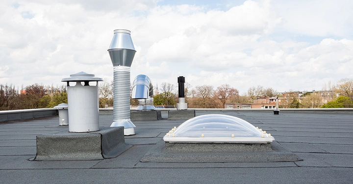 Mehrere Belüftungsschächte und ein Oberlicht befinden sich auf einem Flachdach. Im Hintergrund sind Bäume und Gebäude unter einem bewölkten Himmel zu sehen.