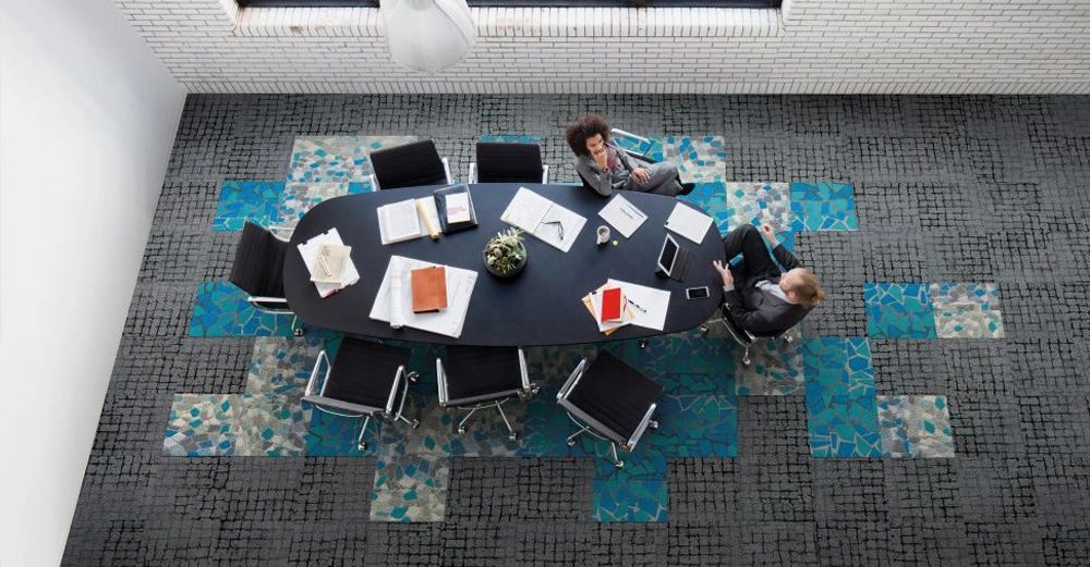 Zwei Personen sitzen an einem großen, ovalen Konferenztisch und arbeiten an Laptops und Dokumenten in einem modernen Besprechungsraum mit gemustertem Teppichboden und weißen Ziegelwänden.