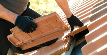 Dachdecker mit schwarzen Handschuhen verlegt an einem sonnigen Tag Tonziegel auf dem Dach.