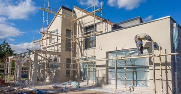 modernes Einfamilienhaus mit Fassadengerüst. Auf dem Gerüst steht ein Maler und verputzt die Außenfassade in Sandfarbe. Blauer Himmel.