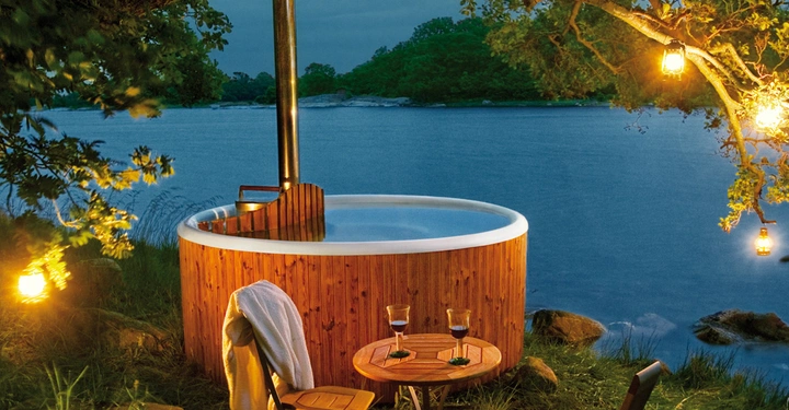 Galerie-Bild 5: Ein runder Holz-Whirlpool steht am Seeufer unter Bäumen mit leuchtenden Hängelaternen ein Tisch und zwei Stühle daneben ein Handtuch auf einem Stuhl im Hintergrund Bäume und Wasser.