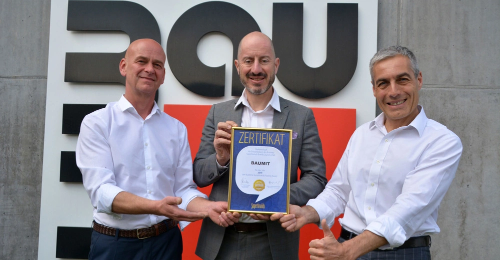 Drei Männer halten ein Zertifikat lächelnd vor einem großen Firmenlogo an einer Betonwand. Text auf dem Zertifikat: "ZERTIFIKAT BAUMIT" accompagné de détails d'accréditation.