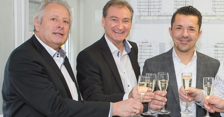 Drei Männer halten Sektgläser und stoßen an. Sie tragen Anzüge und stehen in einem Büro vor einem Planungsdokument an einer Wand.