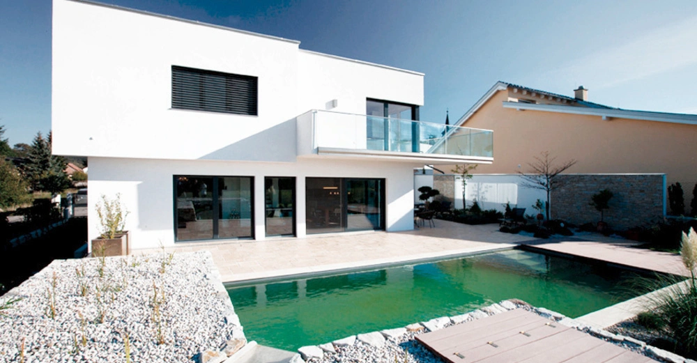 Ein modernes zweistöckiges Haus mit großen Glasfenstern und einem Balkon steht neben einem rechteckigen Swimmingpool in einem gepflegten Garten mit Steinbeeten und einem Holzsteg.