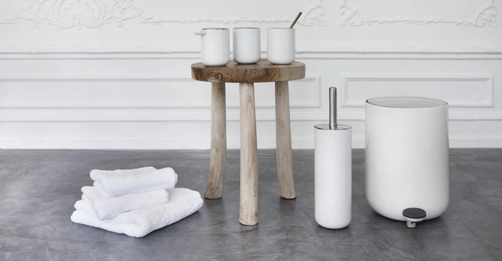 Galerie-Bild 8: Holztisch mit Keramikbehältern und Zahnbürste daneben Wäschetücher auf dem Boden links ein weißer Toilettenbürstenhalter und ein Mülleimer stehen auf einem grauen Boden in einem weiß dekorierten Raum