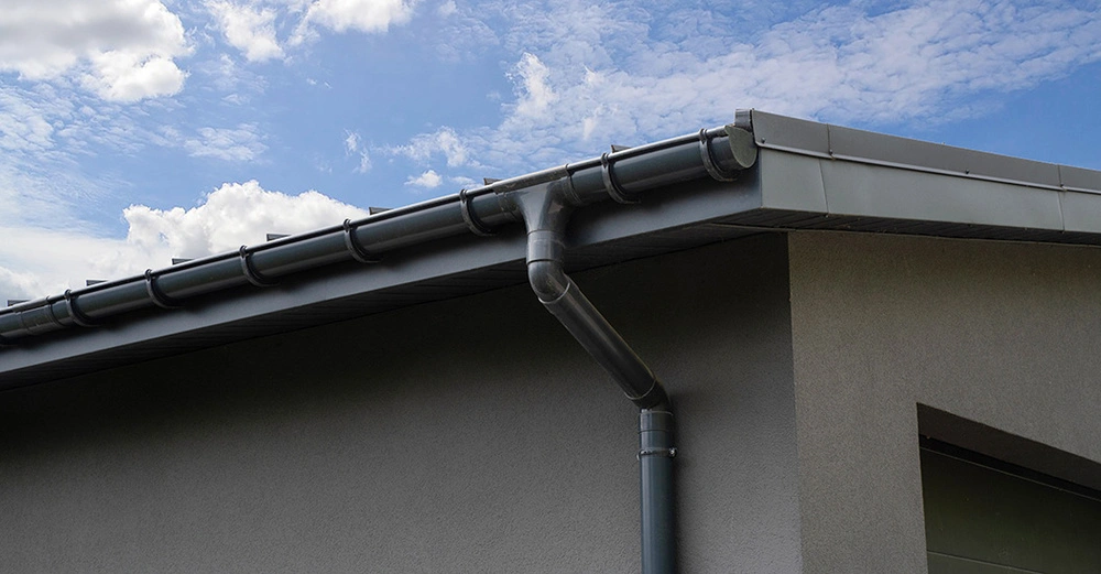 moderne graue Hausfassade mit Dachanschluss und Gesimsadeckungen sowie Regenrinne und Fallrohr in dunklem Anthrazit. 