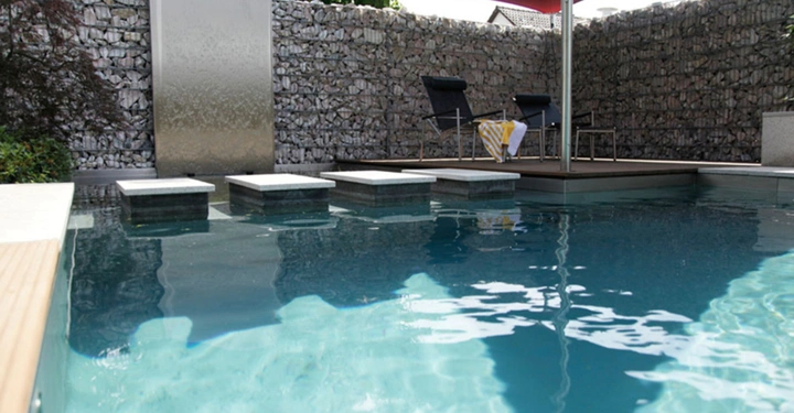 Ein Swimmingpool mit klarem Wasser Steineinfassung und Edelstahl-Wasserfall im Hintergrund Zwei Liegestühle und ein gelbes Handtuch stehen auf einer erhöhten Plattform unter einem roten Sonnensegel.