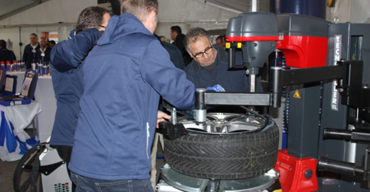 Galerie-Bild 1: Männer arbeiten an einer Maschine zum Reifenwechsel in einem geschäftigen Zelt mit anderen Personen und Ausrüstung im Hintergrund