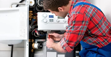 Mann repariert einen Boiler in einem Technikkeller er trägt ein kariertes Hemd und eine blaue Latzhose Werkzeuge und Rohre sind im Hintergrund sichtbar