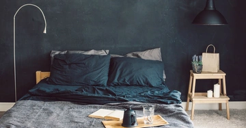 Ein Bett steht in einem modern eingerichteten Schlafzimmer mit dunklen Wänden. Auf dem Bett liegt ein Tablett mit einer Kaffeekanne, einem Glas und einem offenen Buch. Neben dem Bett befindet sich ein Nachttisch mit Pflanzen und Kerzen.