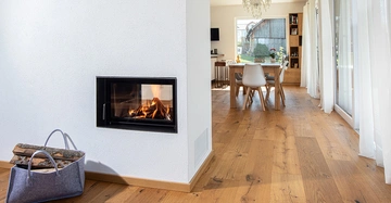 Kaminofen im Einsatz in einem modernen Wohnzimmer mit weiß gestrichenen Wänden und Holzboden. Ein Korb mit Brennholz steht daneben. Im Hintergrund ein Esstisch mit Stühlen und großen Fenstern.